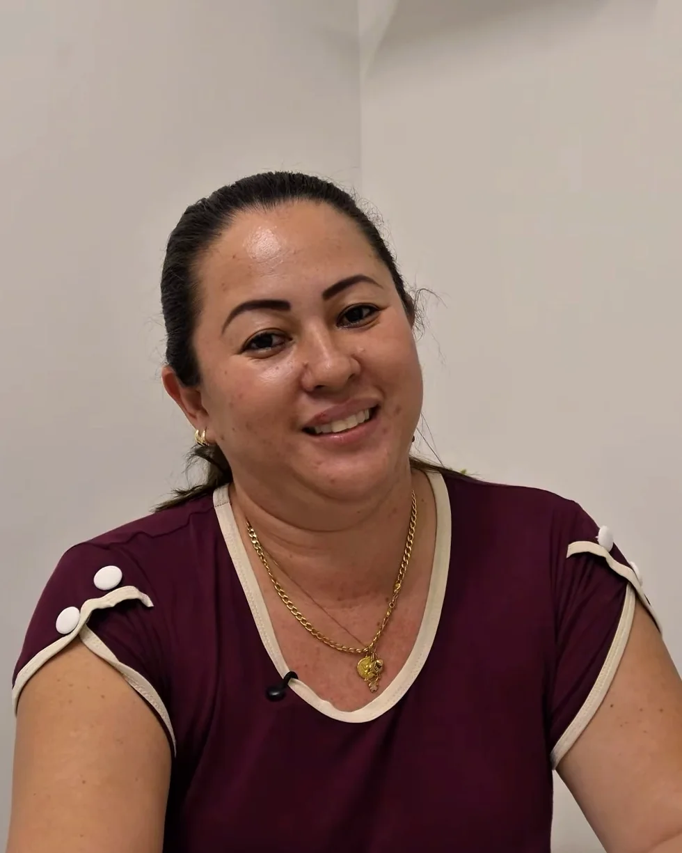 Dra. Telmi sentada à mesa do consultório, de blusa vinho, sorrindo para quem chega