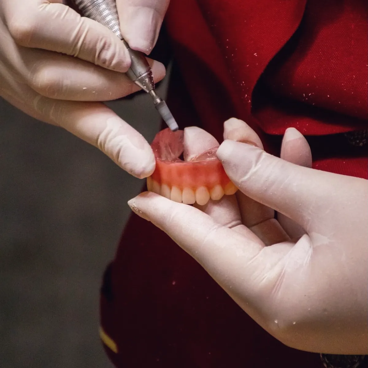 Mãos da Dra. Telmi, de luvas, ajustando uma prótese dentária com a peça de mão; ao fundo, o jaleco vinho de botões dourados.